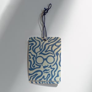 Pode incluir: Ambientador retangular com um design azul e creme. O design apresenta um rosto com óculos e linhas onduladas. Uma corda azul escura está presa no topo. O texto na parte inferior diz: EVITE O CONTACTO COM SUPERFÍCIES PINTADAS, PLÁSTICAS OU ENVERNIZADAS.