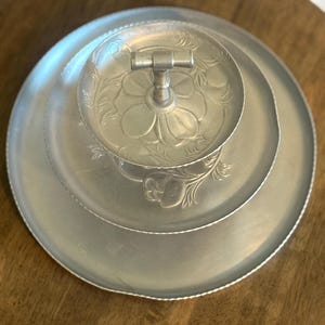 May include: A two-tiered silver-toned metal serving tray featuring a central handle and embossed floral patterns on the circular plates. The tiered stand has a smooth, matte finish with a decorative scalloped edge along the rim of each tray.