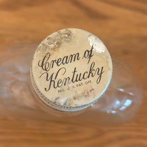 May include: A circular bottle cap with a weathered off-white finish featuring the black cursive text Cream of Kentucky and the smaller printed text REG. U.S. PAT. OFF. The cap sits atop a clear glass bottle with a rounded, smooth neck and body.