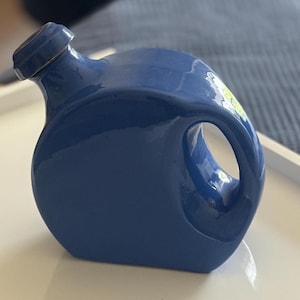 Op de afbeelding: Een glanzend, diepblauw keramieken vat met een cirkelvormig, afgeplat lichaam en een enkele geïntegreerde handgreep. De houder heeft een korte, schuine hals met een bijpassende blauwe dop en een gladde, gelijkmatige afwerking over het geheel.