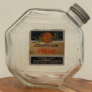 Peut inclure: Une bouteille en verre transparent avec un bouchon à vis métallique incliné et une étiquette vintage indiquant Country Club Brand Pure Apple Cider Vinegar, Full Strength, Contents 1 Quart, Distributed by The Kroger Grocery & Baking Co. Cincinnati, O.