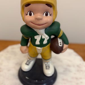 May include: A ceramic figurine of a football player in a yellow helmet and uniform with green accents, holding a brown football. The figure is standing on a black base. The uniform has the number 77.