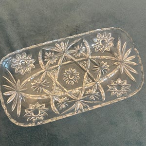 May include: A clear, rectangular glass serving dish with a starburst pattern. The dish features a series of star-shaped and floral designs, creating a decorative and elegant appearance. The edges of the dish are slightly scalloped.