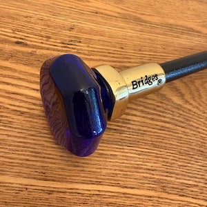 Op de afbeelding: Een close-up van een wandelstok met een donkerblauwe, ovaalvormige handgreep. De handgreep is verbonden met een goudkleurig metalen stuk met de tekst "Bridges". De schacht van de stok is zwart en de achtergrond is een houten oppervlak.