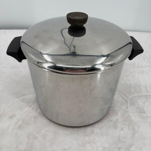 May include: A stainless steel stockpot with a matching lid, featuring two black side handles and a small, round, dark brown knob on top of the lid. The pot has a polished, reflective metallic finish and a cylindrical shape with a flat base.