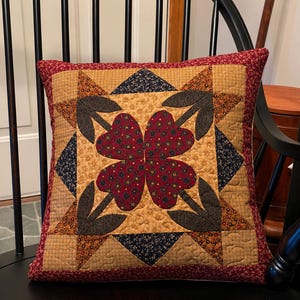 May include: A square quilted throw pillow featuring a central four-leaf clover design made of dark red fabric with small yellow floral patterns. The clover is surrounded by dark gray leaf shapes on a tan background with a red floral border trim.