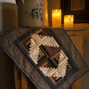 May include: A rectangular quilted table runner featuring a log cabin pattern with various earth-toned fabric squares in shades of brown, tan, cream, and deep red. The runner has a wide, dark gray border with a subtle speckled texture and stitched details.