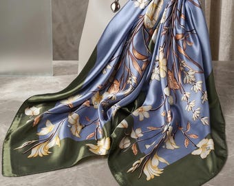以下が含まれることがあります： 花柄のシルクの正方形のスカーフ。スカーフは、クリームと茶色の植物のデザインが施された水色の中心部が特徴です。縁は濃いオリーブグリーンです。スカーフは白いハンドバッグの上にドレープされています。