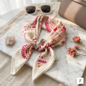 May include: A cream-colored silk scarf with a floral pattern in shades of pink, red, and green. The scarf is knotted and displayed on a marble surface, with sunglasses and a purse in the background. The scarf has a square shape.