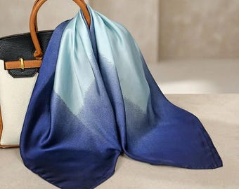 以下が含まれることがあります： ライトブルーからネイビーへのグラデーションデザインの正方形シルクスカーフ。 黒と白のハンドバッグにかけられており、茶色のハンドルが付いています。 スカーフの滑らかな質感と配色から、ファッションアクセサリーであることがわかります。