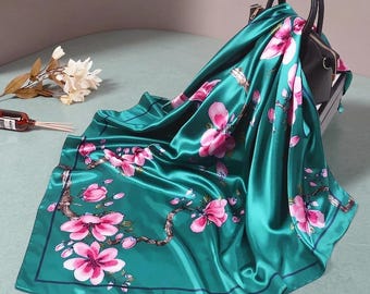 以下が含まれることがあります： ピンクの花と茶色の枝のフローラルパターンが特徴の、ティールシルクのスカーフ。スカーフは黒いハンドバッグの上に掛けられています。スカーフは濃い青色の縁取りと光沢のある滑らかな質感です。