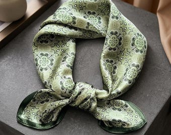 以下が含まれることがあります： 緑色のシルクのスカーフ。緑と黒の色合いで花柄が繰り返されています。スカーフは結ばれており、端が広がっています。スカーフには濃い緑色の縁取りがあります。