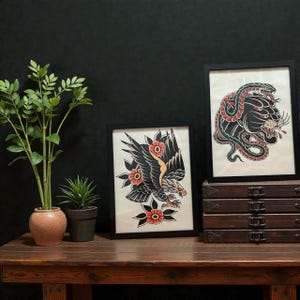 Puede incluir: Dos impresiones artísticas enmarcadas al estilo tatuaje. Una presenta un águila negra con detalles florales naranjas y rojos. La otra representa una pantera negra entrelazada con una serpiente verde, con detalles rojos. Los marcos son negros.