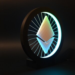 May include: A black, circular LED light featuring a geometric Ethereum symbol. The symbol is white and has a multi-coloured glow. The light has a stand and a power cable.