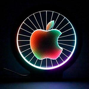 Könnte beinhalten: Leuchtendes Apple-Logo in einem kreisförmigen Rahmen mit radialem Muster. Der Apfel hat einen Farbverlauf von Rot, Grün und Gelb vor dunklem Hintergrund. Rahmen und Logo sind in verschiedenen Farben beleuchtet.