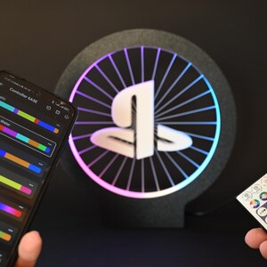 May include: A black and gray LED light with a white logo and a colorful light ring. A smartphone displays a color control interface. A remote control is also visible. The light is designed for gaming.