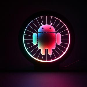 May include: A colour-changing Android logo light. The logo is in the centre of a black circular frame with a radial pattern. The light transitions through a spectrum of colours, including red, green, and blue, against a dark background.