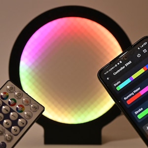 Pode incluir: Uma luz LED circular preta com um ecrã colorido e pixelizado. Um controlo remoto e um smartphone que exibe um painel de controlo estão em primeiro plano. O ecrã da luz alterna entre várias cores, criando um efeito vibrante.