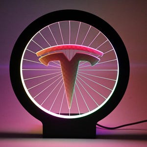 May include: A black, circular LED lamp featuring the Tesla logo. The logo is a stylized 'T' shape, with a gradient of colours. The lamp has a wheel-like design with radiating lines and a black base.