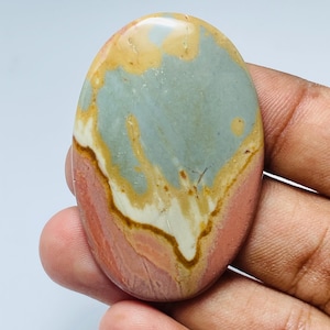 May include: An oval polished stone with a mix of colours. The stone has a light blue-grey centre, edged with tan and white, and framed by a reddish-pink border. The stone is held in a hand.
