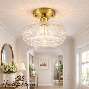 May include: A flush mount ceiling light fixture with a gold-toned base and a clear, patterned glass shade. The shade has a floral design. The light is installed in a hallway with white walls and trim.