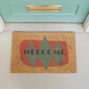 May include: A rectangular coir doormat with a retro design. The mat features a red and green geometric pattern with the word "WELCOME" in black. The mat is placed in front of a teal door with a gold mail slot.