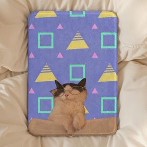Op de afbeelding: Een rechthoekig, blauw kattenbed met een geometrisch patroon van gele piramides, roze driehoeken en groene vierkanten. Een kat met een bril staat centraal op het bed. Het bed ligt op een wit oppervlak.