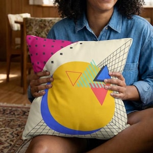 May include: A cushion with a retro geometric design. The cushion features a yellow circle, blue semi-circle, pink square, and a grid pattern. The cushion is cream-coloured with a variety of shapes and colours.