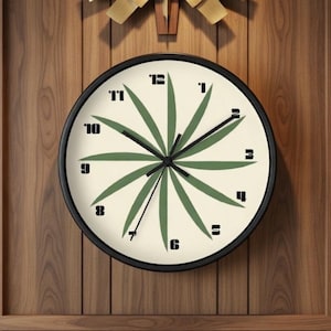 May include: A round wall clock with a black frame, featuring a cream-colored face. The clock face has green leaf-like designs radiating from the center, with black numerals marking the hours. The clock is mounted on a wooden wall.