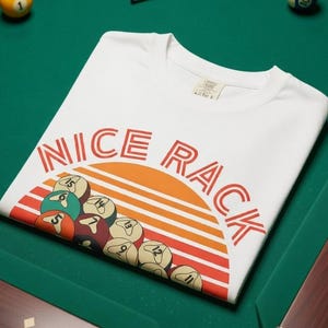 Puede incluir: Camiseta blanca con el texto "NICE RACK" en rojo, sobre un gráfico de bolas de billar y un diseño de puesta de sol. La camiseta está sobre una mesa de billar verde, con bolas de billar y un taco.