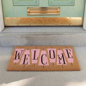 Pode incluir: Um tapete retangular com a palavra "WELCOME" em letras pretas e formas recortadas rosa. O tapete é feito de um material de fibra natural e é colocado em frente a uma porta verde menta.