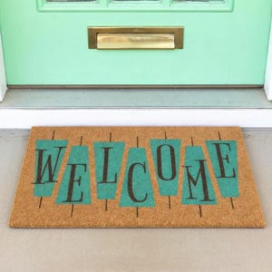 Pode incluir: Um capacho retangular feito de fibra de coco natural. O tapete apresenta a palavra "WELCOME" em letras maiúsculas verde-escuras com detalhes retangulares turquesa. O tapete é colocado em frente a uma porta verde-menta.