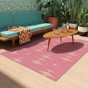 May include: A mid-century modern outdoor patio scene featuring a pink rug with a geometric pattern, a wooden coffee table, and a teal sofa with a patterned throw. Potted plants and a decorative wall complete the space.