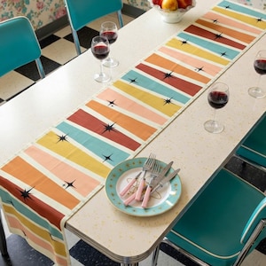 May include: A retro-style table setting featuring a colorful striped table runner with starburst accents. The runner is placed on a white table, complemented by teal chairs, wine glasses, and a plate with pink-handled silverware. A bowl of apples adds a touch of freshness.
