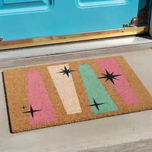 Op de afbeelding: Een rechthoekige deurmat van natuurlijke kokosvezel. De mat heeft een retro-ontwerp met roze, gebroken witte en groene verticale strepen, elk versierd met een zwarte ster. De deurmat ligt voor een turquoise deur.