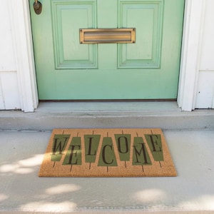 May include: A rectangular doormat with the word "WELCOME" in dark green block letters. The mat is made of a natural fiber material and is placed in front of a light green door.