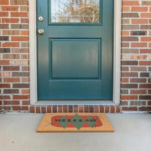 May include: A rectangular doormat with the word "WELCOME" in a retro font. The mat has a tan background with green and red diamond shapes. The mat is in front of a teal door and a brick wall.