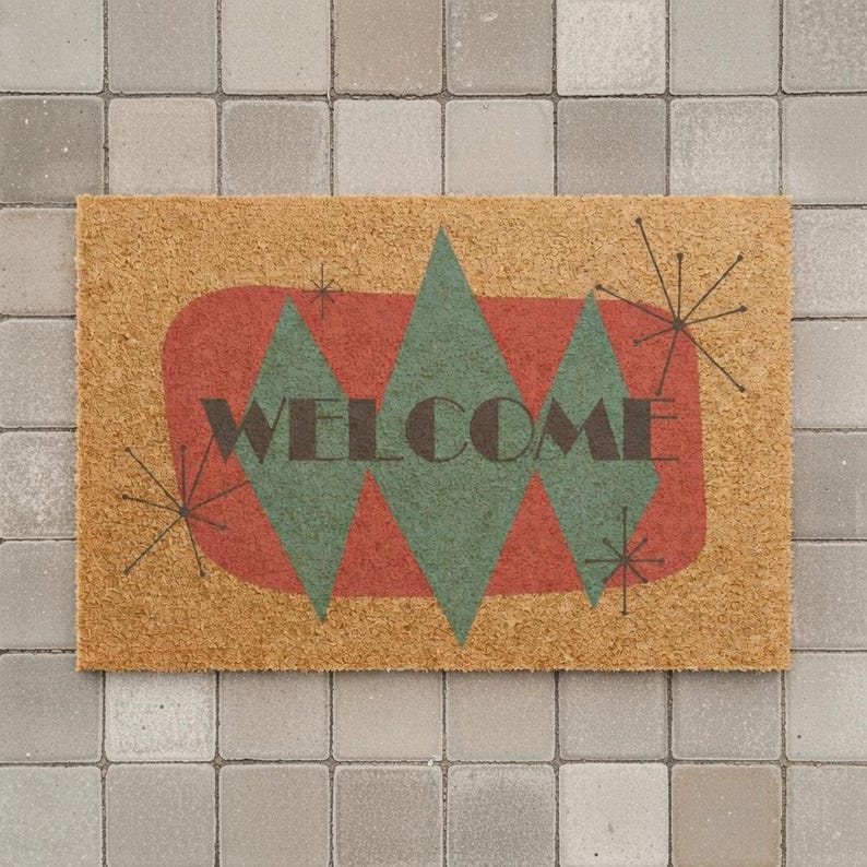 May include: A rectangular coir doormat with a retro design. The mat features a red oval with three green diamond shapes and the word "WELCOME" in dark brown. Black starburst designs are on the corners.