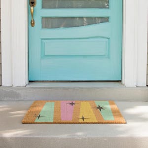 May include: A rectangular doormat with a retro design. The mat features vertical stripes in pastel green, pink, and yellow, with black starburst accents. The mat is made of a natural fiber material and is placed in front of a turquoise door.