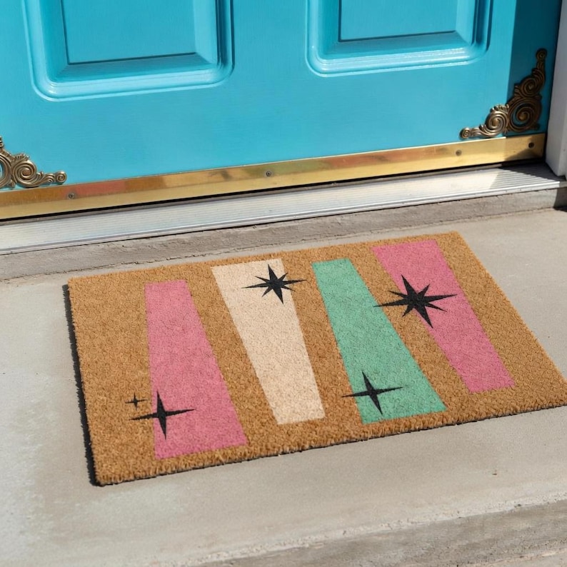 Op de afbeelding: Een rechthoekige deurmat van natuurlijke kokosvezel. De mat heeft een retro-ontwerp met roze, gebroken witte en groene verticale strepen, elk versierd met een zwarte ster. De deurmat ligt voor een turquoise deur.