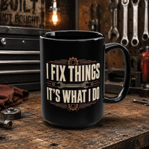 May include: A black ceramic mug featuring a graphic design with the text I FIX THINGS IT'S WHAT I DO in a beige font. The design includes a central wrench illustration and gear motifs above and below the text, all centered on the mug's front.