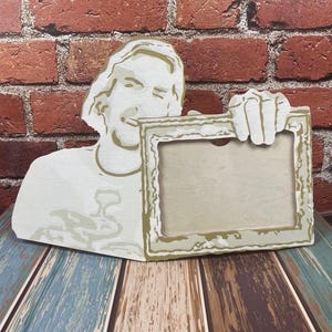 May include: A decorative wooden photo frame featuring a stylized image of a person holding the frame. The frame has a light beige color with gold accents and a distressed edge. The background is a brick wall and a wooden surface.