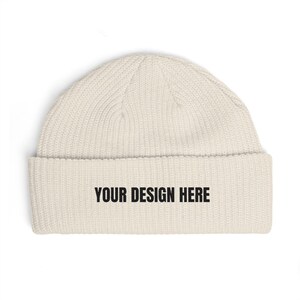 May include: Cream-colored beanie hat with a ribbed knit texture. The folded brim features the text "YOUR DESIGN HERE" in bold black letters. The hat is a winter accessory.