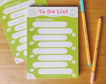 Może przedstawiać: Zielony notes do listy rzeczy do zrobienia z motywem żaby i królika. Notes ma białe tło z zielonymi chmurkami do wpisywania zadań. Tekst "To Do List with frog and bun" jest nadrukowany na górze notesu.
