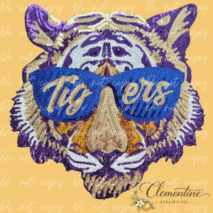 May include: A sequined tiger head patch with a blue pair of sunglasses that read "Tigers". The tiger is in shades of purple, gold, and white. The patch is likely for apparel or accessories.