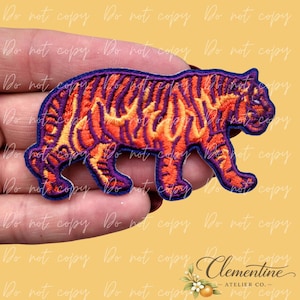 Può includere: Toppa ricamata a forma di tigre con strisce arancioni e viola a forma di fiamma, bordate di viola. La toppa è in posa da camminata. Il testo "Do not copy" è ripetuto sullo sfondo.