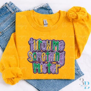 May include: A yellow sweatshirt with the phrase "Throw Me Something Mister" in purple, green, and yellow lettering. The sweatshirt is folded on top of a pair of blue jeans.