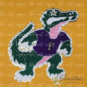 May include: A patch featuring a green alligator mascot wearing a purple shirt with a gold letter F on the chest. The alligator has a pink tongue, a gold star on its head, and a shimmering gold glitter border surrounding the entire design.