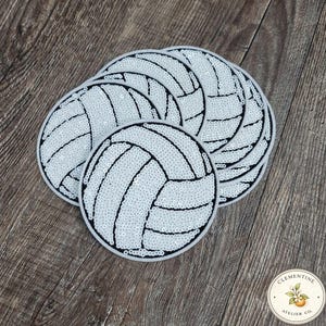 Könnte beinhalten: Eine Sammlung weißer, paillettenbesetzter Untersetzer in Volleyballform mit schwarzen Details. Die Untersetzer sind auf einer Holzoberfläche angeordnet und zeigen ihre runde Form und ihr strukturiertes Design. Das Bild enthält ein Logo in der unteren rechten Ecke.
