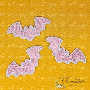 Puede incluir: Tres parches bordados de murciélagos en color rosa claro con un borde blanco. Cada murciélago se muestra con las alas extendidas en posición de vuelo. Los parches están dispuestos sobre un fondo de color amarillo mostaza sólido.